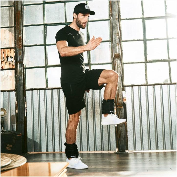 Uomo in posa fitness con cavigliere Gorilla Sports, abbigliamento sportivo nero, interno industriale